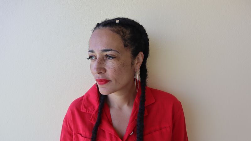 Wearing a bright red shirt, a woman with braided hair and freckles stands looking towards her side.