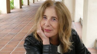 A woman sits at a table with her head in her hand, staring into the camera with a warm smile.