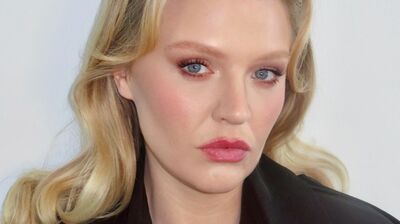 A woman with wavy blonde hair and blue eyes poses for the camera, wearing a black coat against a plain background.