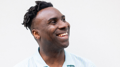 A man smiles and claps his hands together, wearing a light blue shirt against a plain background.