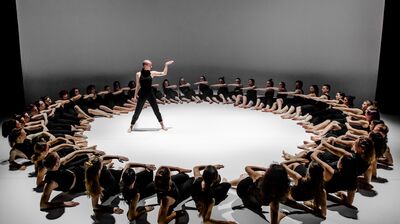 Large group of dancers lying down in a circle around one stood dancer lifting one arm and look at their hand