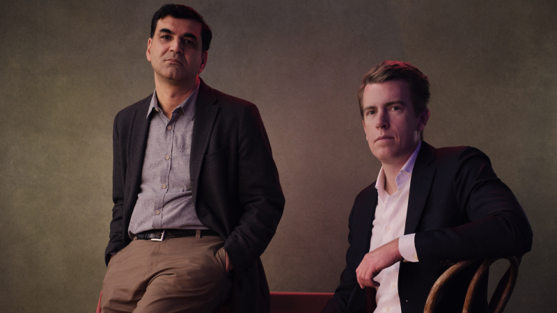Hosts of The Rest is Classified sitting at a table wearing suits against a grey background.