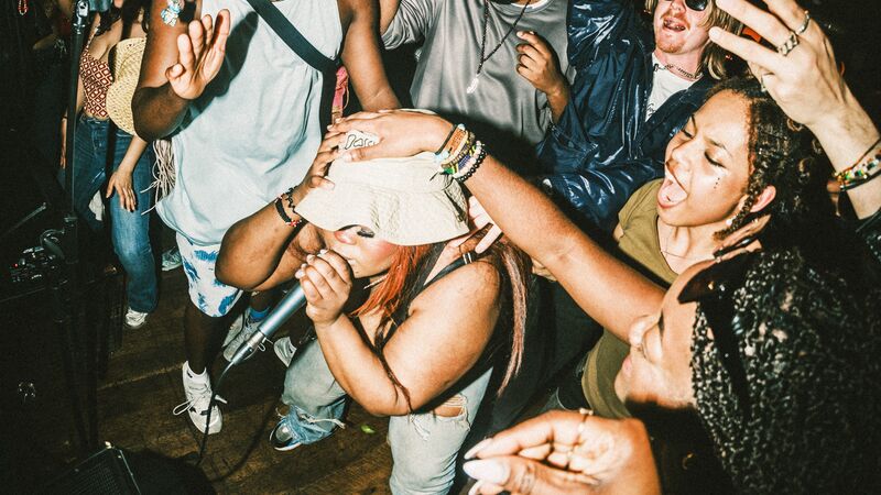 A female performer wearing a bucket hat sings into a microphone amidst a dense, joyful crowd at a lively concert or party, with a man in a light blue furry hat dancing behind her.