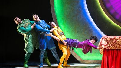 Four brightly costumed acrobats or actors, in a human chain, strain dramatically against a low red table and a backdrop of large, colorful glowing arches on a stage.