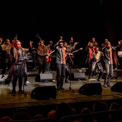 A large African music ensemble performs on a stage, with singers and instrumentalists wearing vibrant traditional clothing featuring colourful neck collars and beadwork