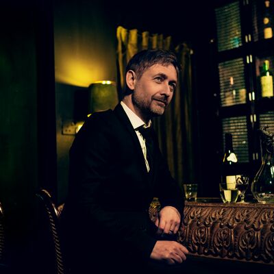 A man in a suit sits at a dark bar looking away from the camera.