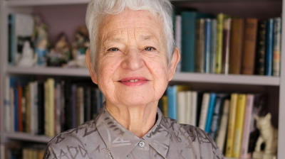 An author stood in front of a book case wearing a grey patterned shirt