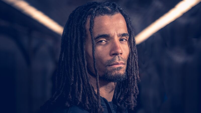 Close-up of a man with long dreadlocks and a beard, looking at the camera.