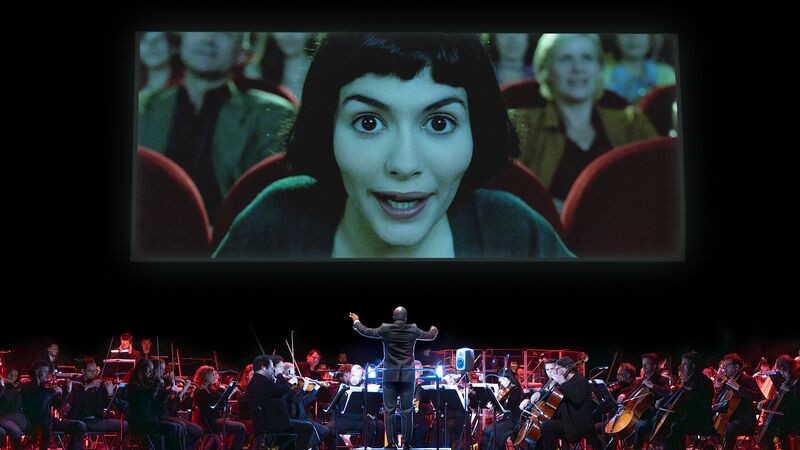 A stage showing a symphony orchestra playing below a large screen displaying a close-up of a woman from a film, looking intently at the audience