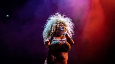 Midgitte Bardot stands in a fishnet dress and green bra with bright blonde hair that fans out against the purple and red lit backdrop.