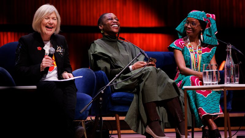 Three people on stage sitting on blue chairs against a red background.