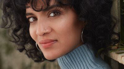 Close-up of a woman with curly dark hair and a nose ring, wearing a blue turtleneck in front of green foliage.