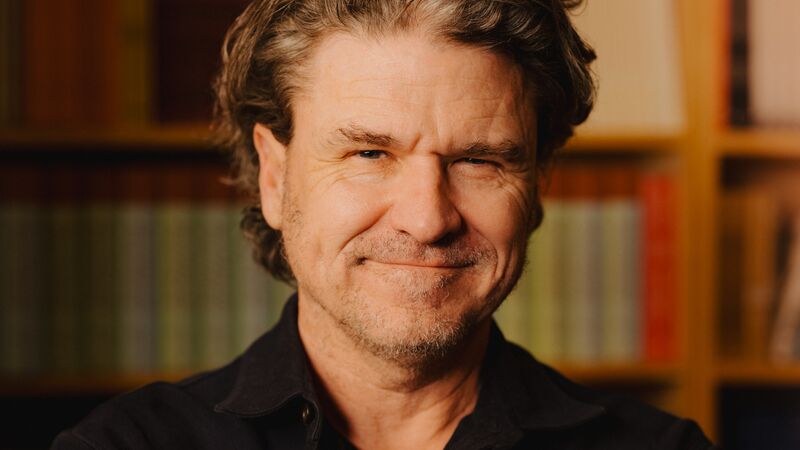 Dave Eggers wearing a black shirt standing in front of a bookshelf.