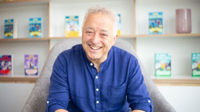Frank Cottrell Boyce wearing a blue shirt sitting in front of a shelf of books.