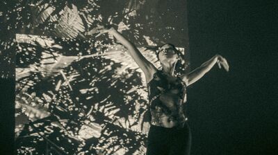 A dancer performs with arms outstretched against a backdrop of grainy, black-and-white projected leaf patterns.