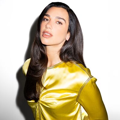 Dua Lipa with long straight brown hair, wearing a satin yellow shirt.