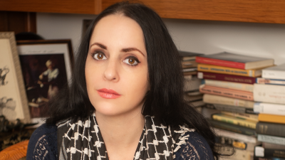 Molly Crabapple sitting writing at a desk in front of a bookcase.