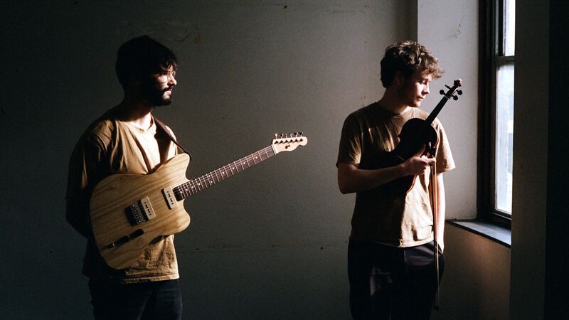 Two young musicians hold a natural wood electric guitar and a violin in a stark room lit by window light.
