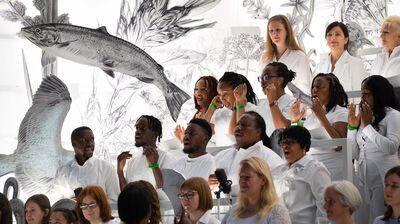 A diverse choir in white clothing sings passionately in front of a giant black and white sketch of a fish and flora.