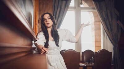 A woman in a white puff-sleeve dress gestures dramatically while leaning on wood paneling inside a historic room.