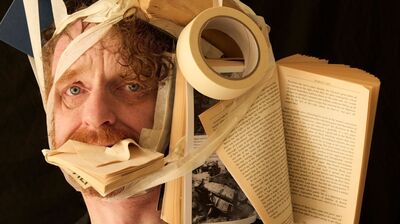 a man in a black blazer looking directly at the camera while a chaotic cluster of books and a roll of masking tape are bound tightly to his head and stuffed into his mouth using beige tape.