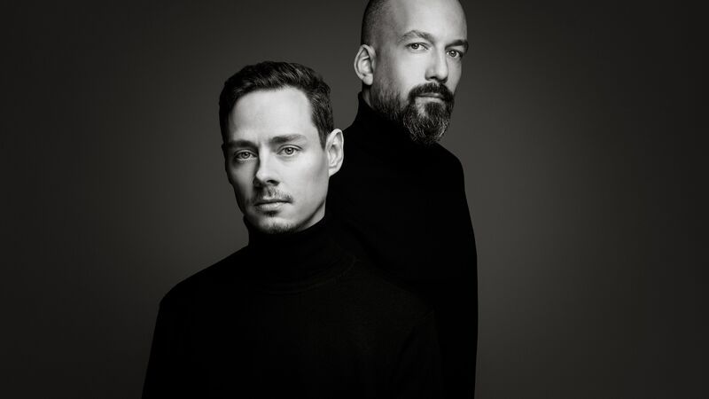 A black-and-white studio portrait of two men in black turtlenecks standing staggered against a grey gradient background.