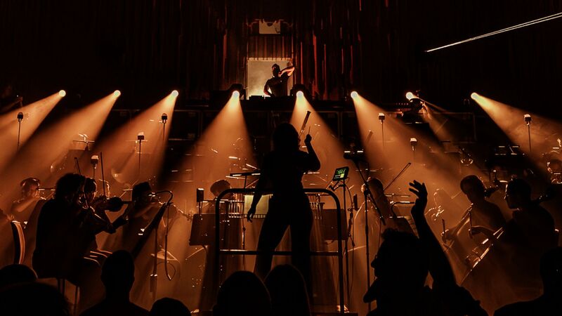 Lights shine out from a dark stage, showing the silhouette of an orchestra.
