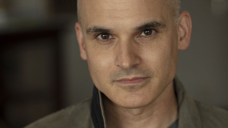 A close-up portrait of a man with short buzzed hair looking into the camera, wearing an olive-green buttoned jacket.