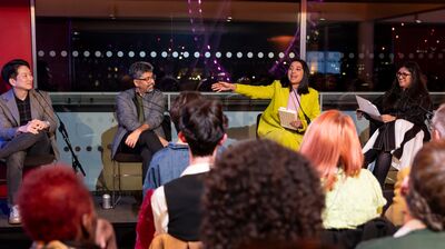 Four panelists sit on a stage speaking into microphones before an audience, with large windows overlooking a city at night