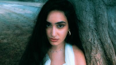 A woman with long dark hair and a light-colored top leans against a tree trunk in an outdoor setting.