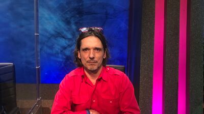 Jeremy Deller in a bright pink shirt, sat behind a lit up sign with his name and a quiz buzzer.