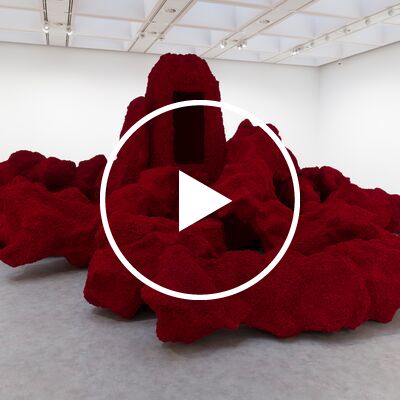 A large, textured dark-red sculpture resembling an organic rock formation sits in the center of a bright, white-walled museum gallery, featuring a tall central monolith with a void-like black rectangular opening.