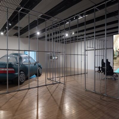 A view of the exhibition Musquiqui Chihying: A Life of Navigation in the Hayward Gallery's HENI Project Space. Vertical metal poles divide the space; to the left hand side we see the inflatable work Toyota – an inflatable car; and to the right a screen displays another video work.