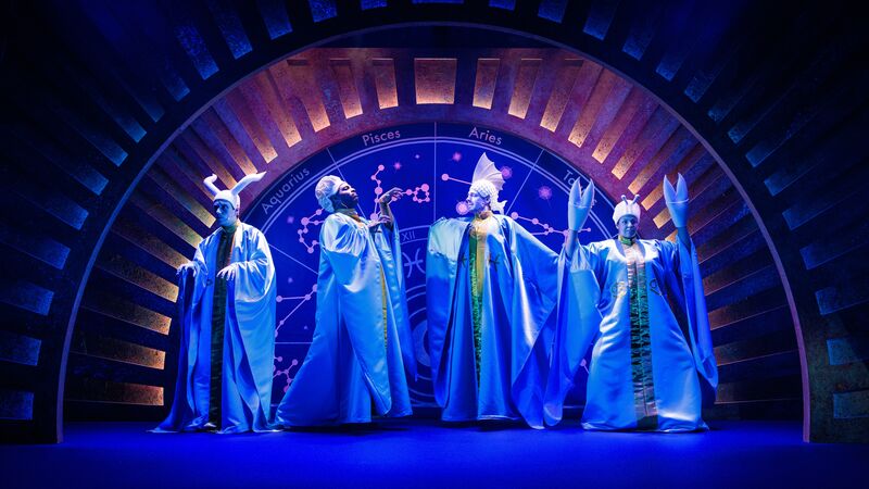 Four actors dressed as gods stand on a stage in front of a zodiac wheel, under blue lighting