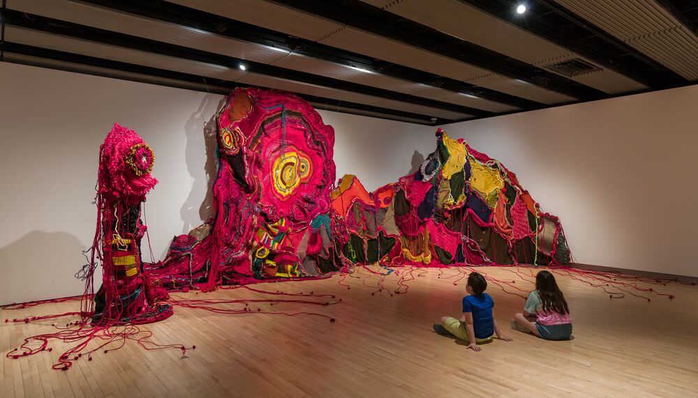 Installation view of Aluaiy Kaumakan with two children sitting in front of artwork