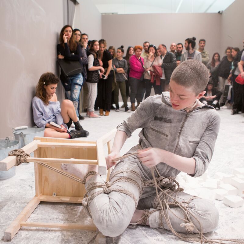 Performance artist binding their legs to a chair in front of an audience in a white room.