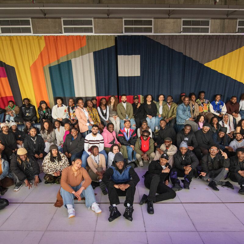 Large group of young people of colour sitting in front of a tapestry in the Queen Elizabeth Hall.