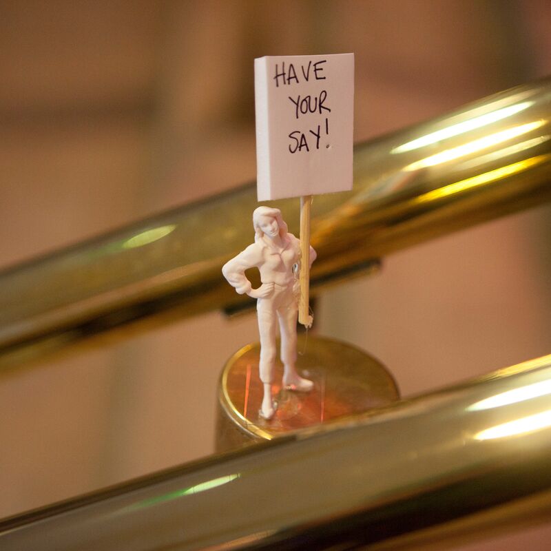 A small plastic figurine of a woman holding a sign, which says "Have Your Say!" It sits on a brass balustrade