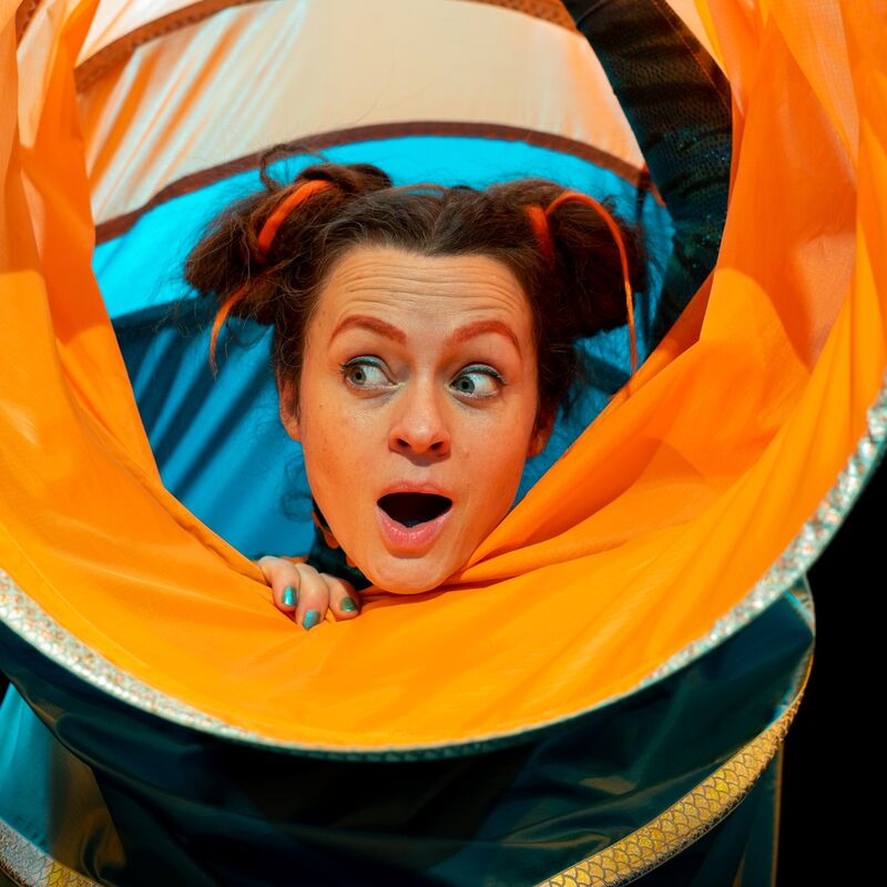 Actor inside a colourful fabric tube looking to the left with their mouth open.