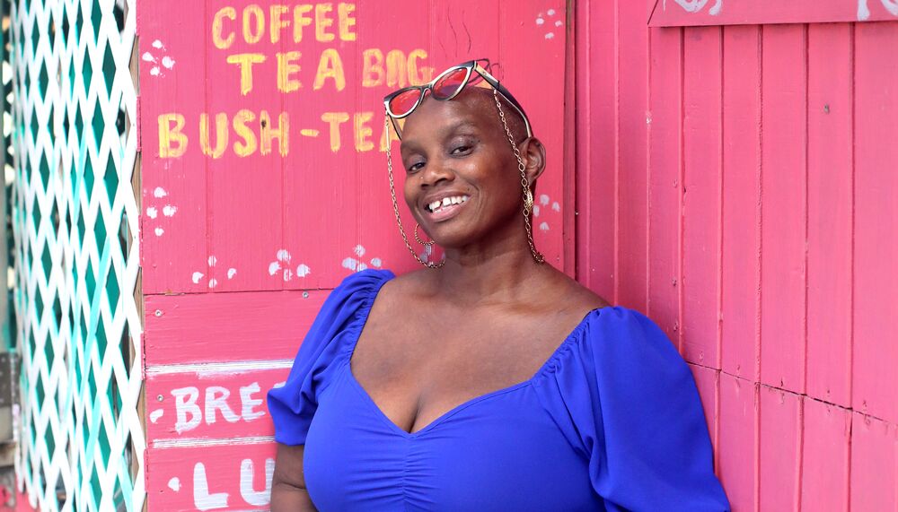 Andi Oliver leans against a pink food shack wearing a blue v-necked dress. She wears her glasses on her head and smiles to camera.