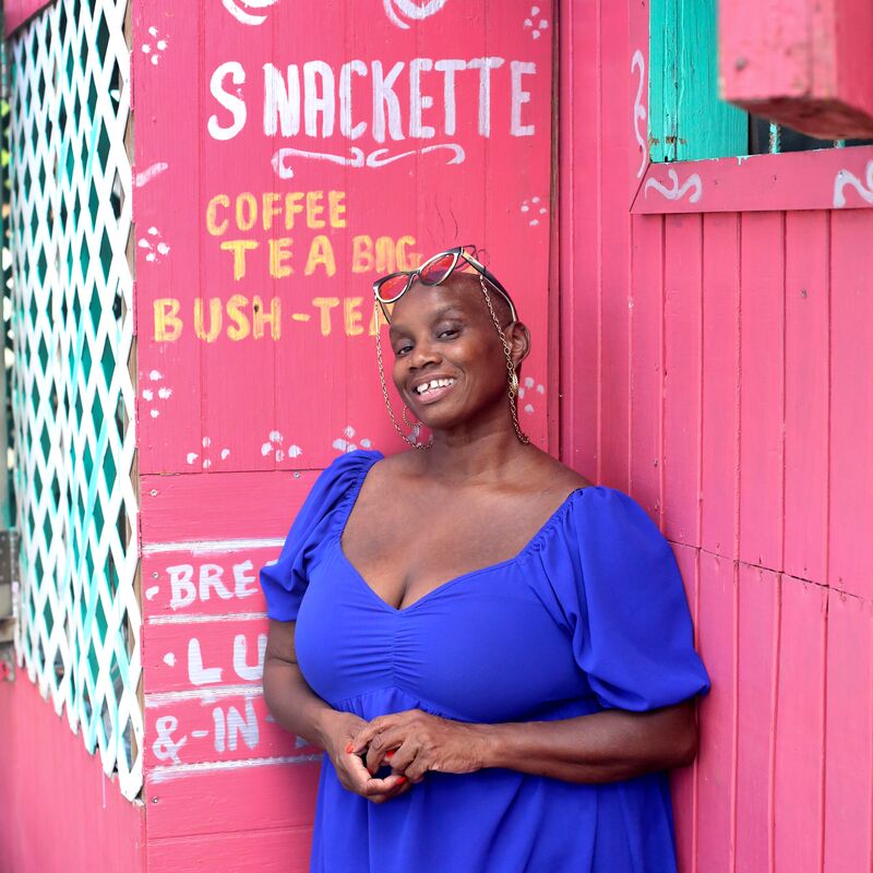 Andi Oliver leans against a pink food shack wearing a blue v-necked dress. She wears her glasses on her head and smiles to camera.