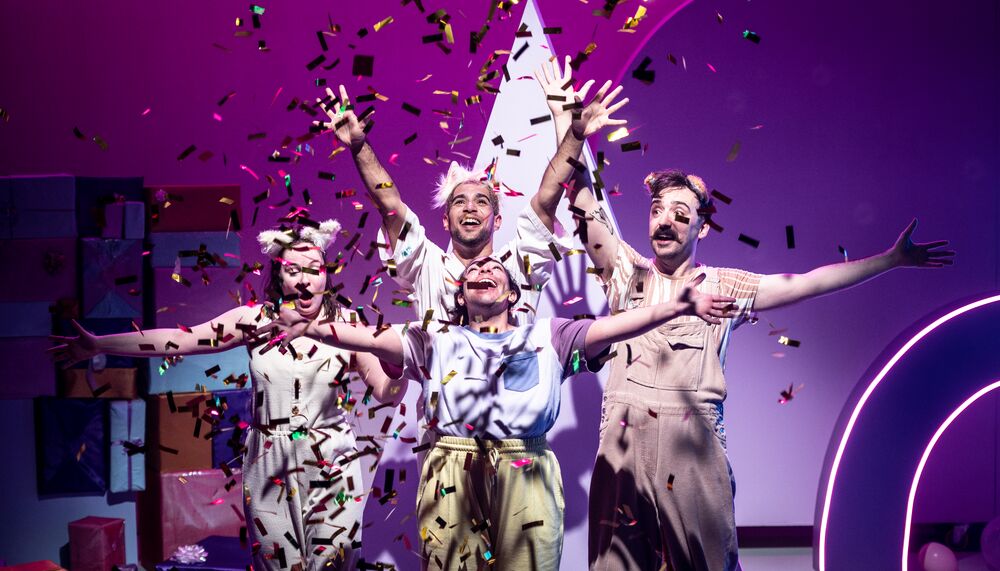 4 performers hold their arms up in celebration with smiles on their faces surrounded by floating metallic confetti.