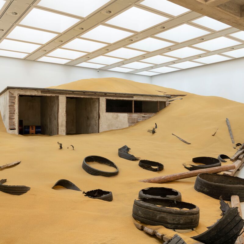 Installation shot of the Mike Nelson exhibition, showing a ruined building half covered in sand with debris strewn about
