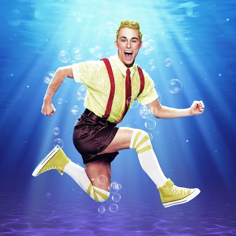 A man with blonde hair is posing mid-air in a running motion against an underwater backdrop. He wears yellow trainers and knee-high white socks with brown shorts and red braces over a white shirt. He is looking excitedly at the camera.