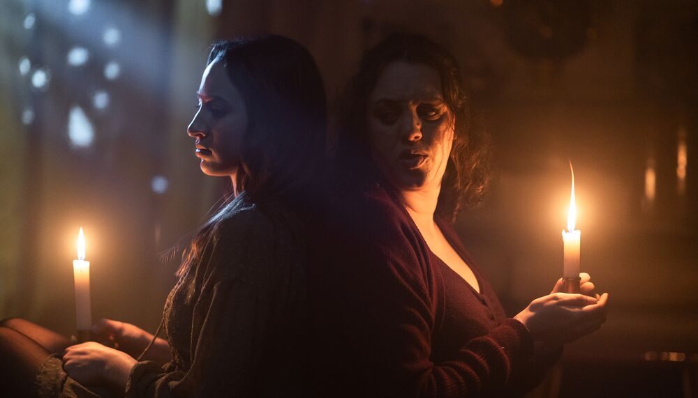 Two women sit back to back in the dark. They hold candles which light their faces.