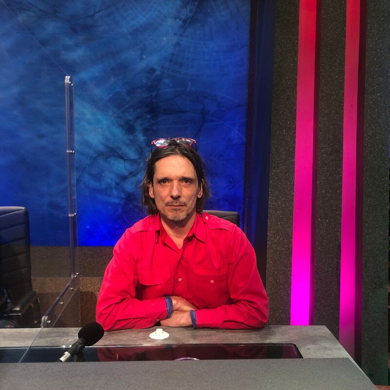 Jeremy Deller wears a bright red shirt, sat at a desk
