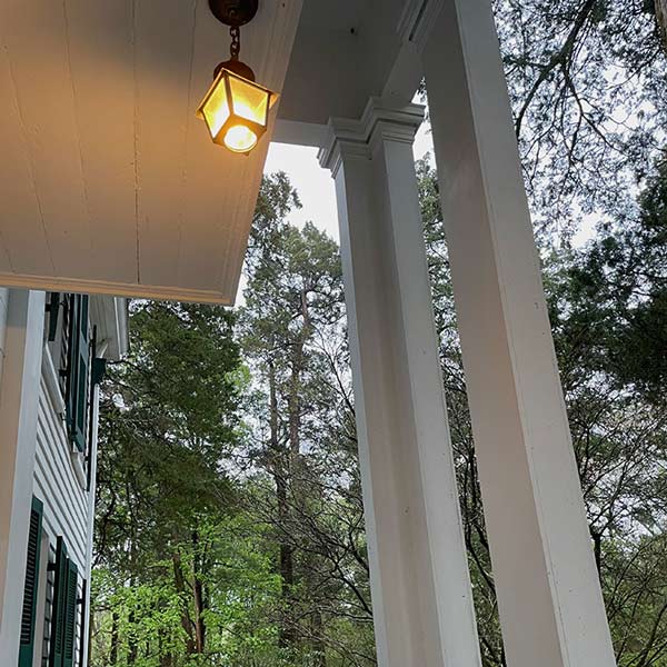Photo taken right outside front door of Rowan Oak. Porch ceiling, hanging lantern light, and porch columns comprise most of the foreground. Trees and side of home make up the background.