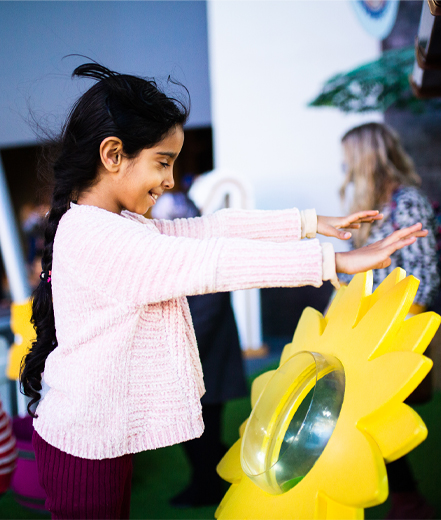 Photo of a girl smiling, with her arms lifted up to shoulder level and air blowing through her hair. She's in front of a giant yellow sunflower exhibit