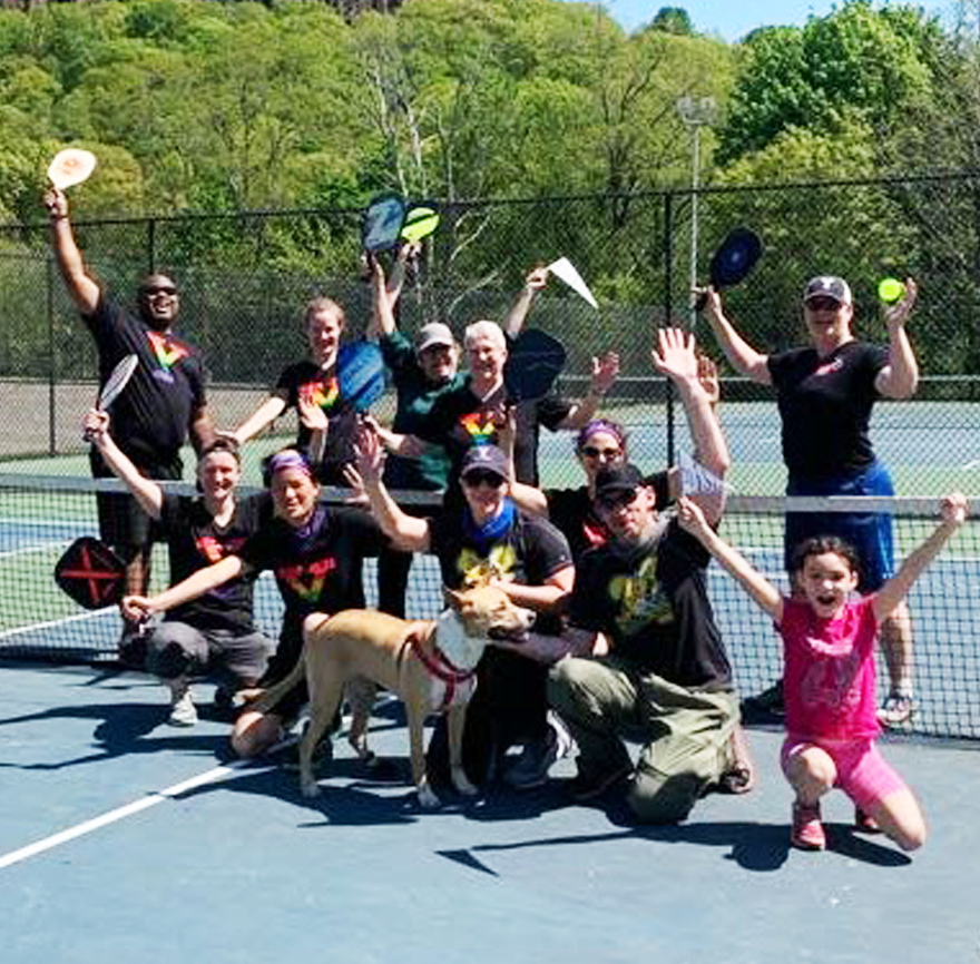 a cheering group of humans and one dog with pickleball paddles on a junior tennis court.